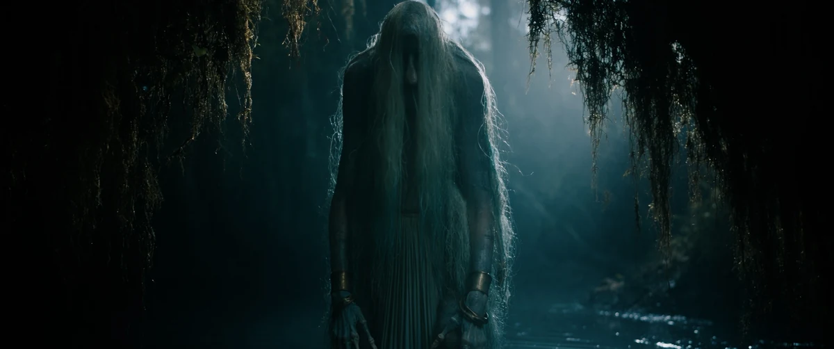 A tall veiled figure stands in a misty forest pool under hanging moss