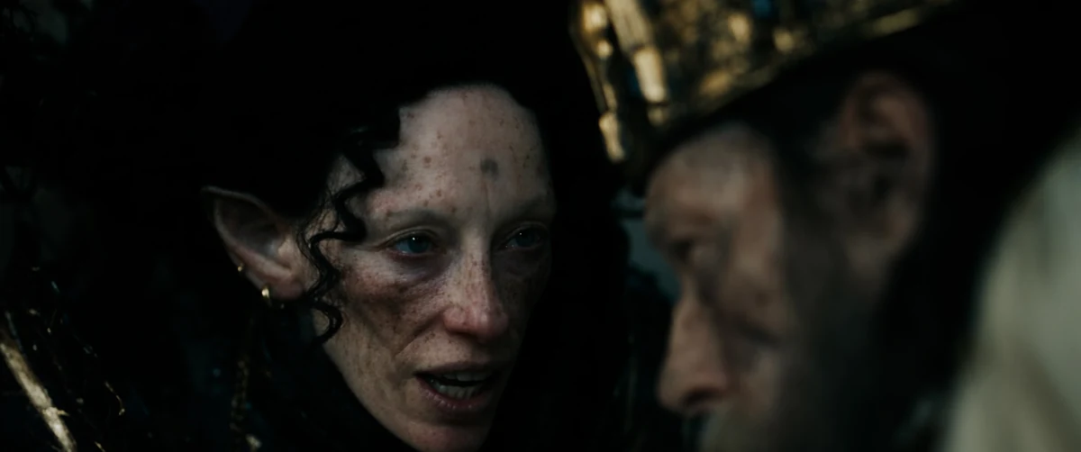 An elfin woman leans close to a crowned, bearded king in candlelit darkness