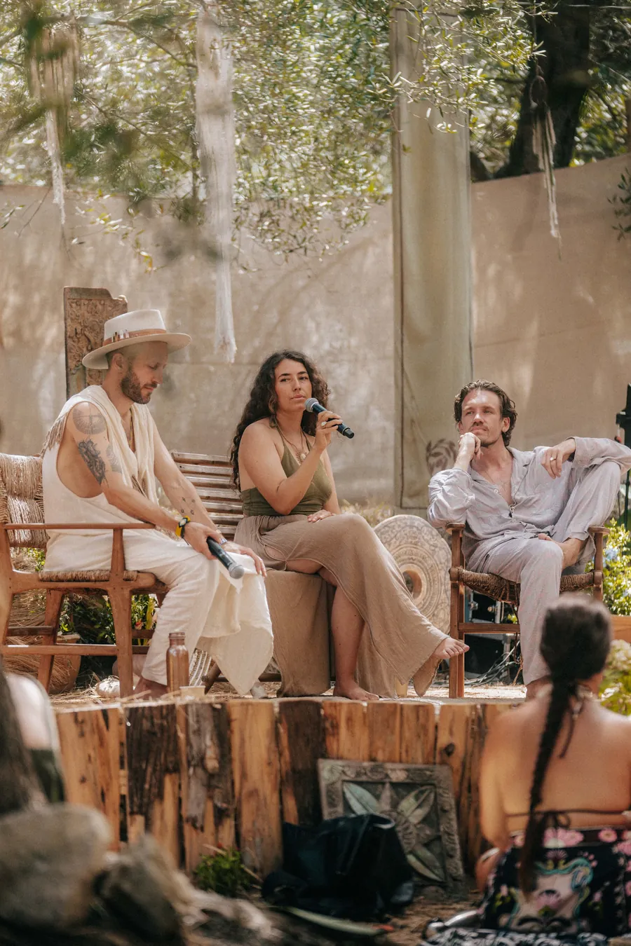 Three speakers in conversation on a wooden stage among olive trees
