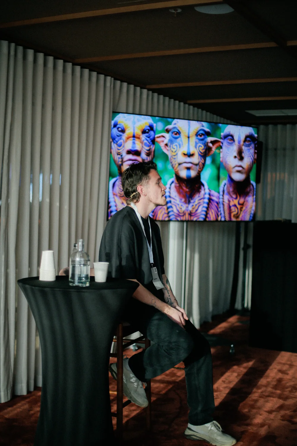 David seated at a high table beside a screen showing three painted creature faces