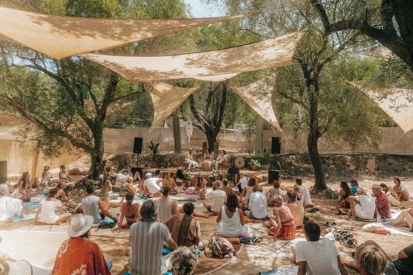 An audience seated on rugs in an olive grove under sun sails, facing a small stage.